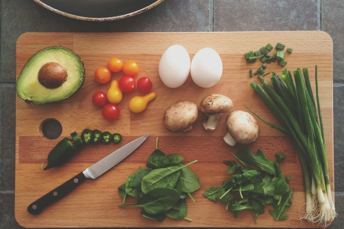 A knife, avocado, eggs, and vegetables arranged on a wooden cutting board.
