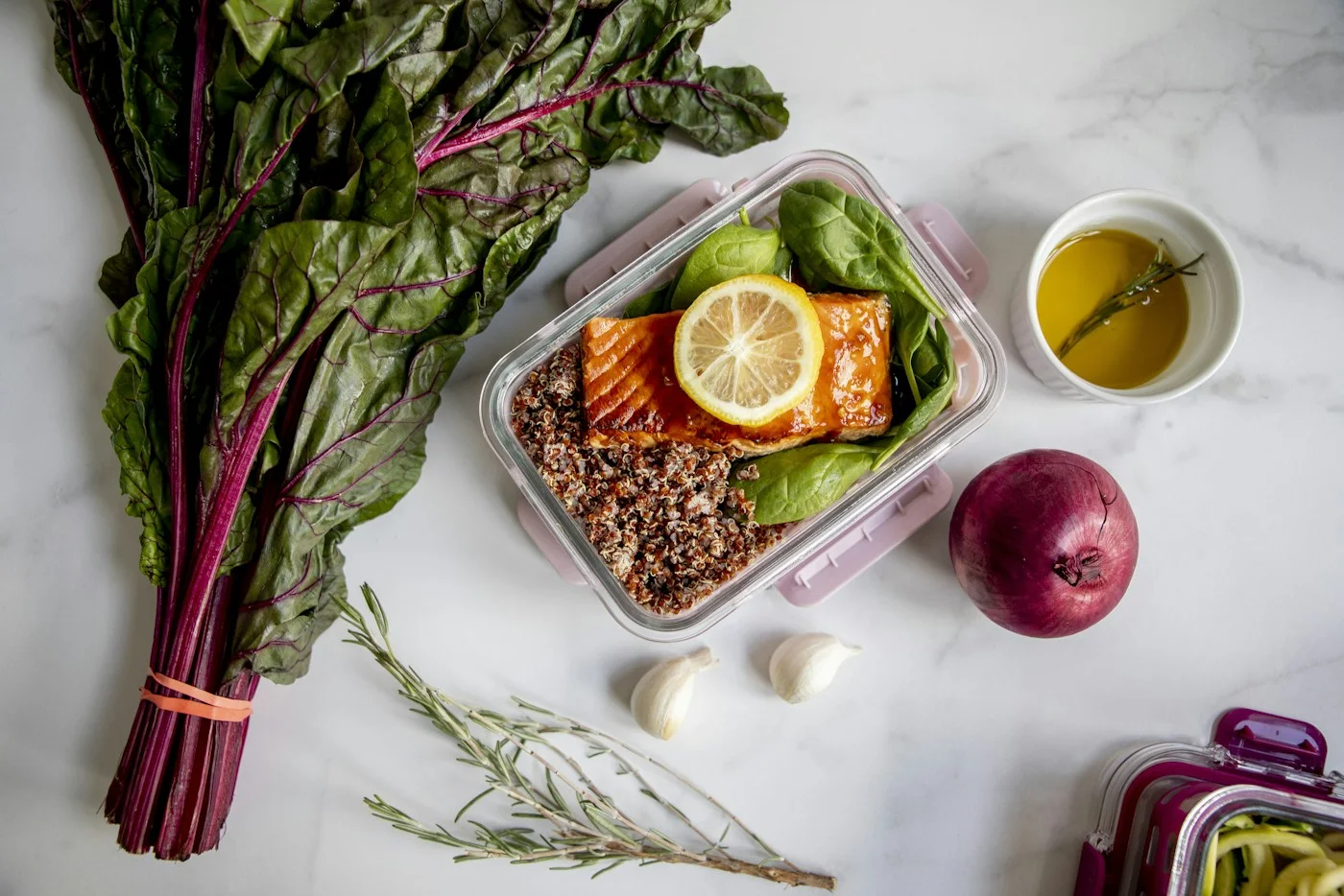 A glass lunch container filled with grains, greens, and lemon beside fresh vegetables.