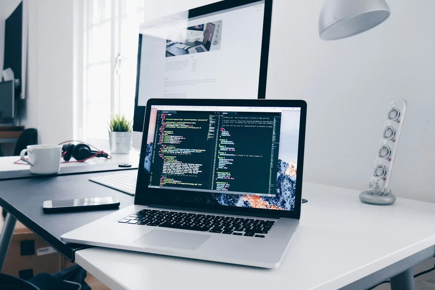 A laptop and monitor on a white desk near a bright window.